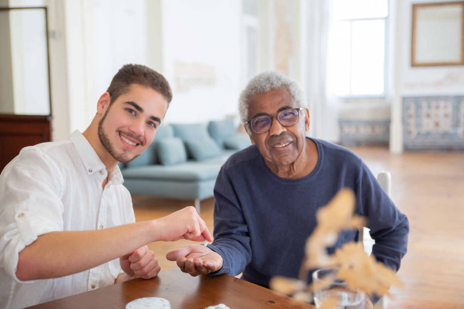 A young carer helps an older man with his medicines at the kitchen table.